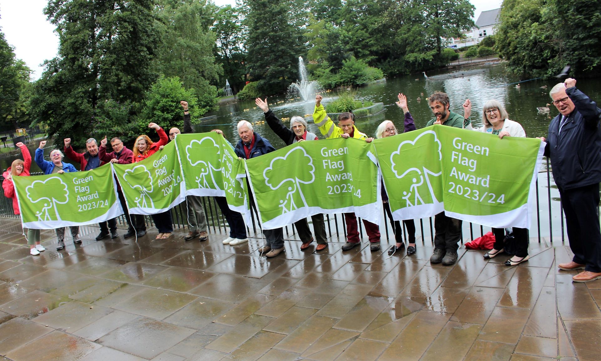 A large group of people stood in a line holding 5 seperate Green Flag Award 2023 flags. The group are stoof in a park in front of a lake.